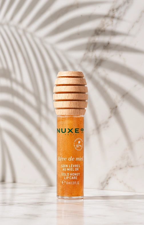 Envase del Tratamiento Labial con Miel Gold de NUXE con tapón de madera, sobre mármol con sombra de palmera
